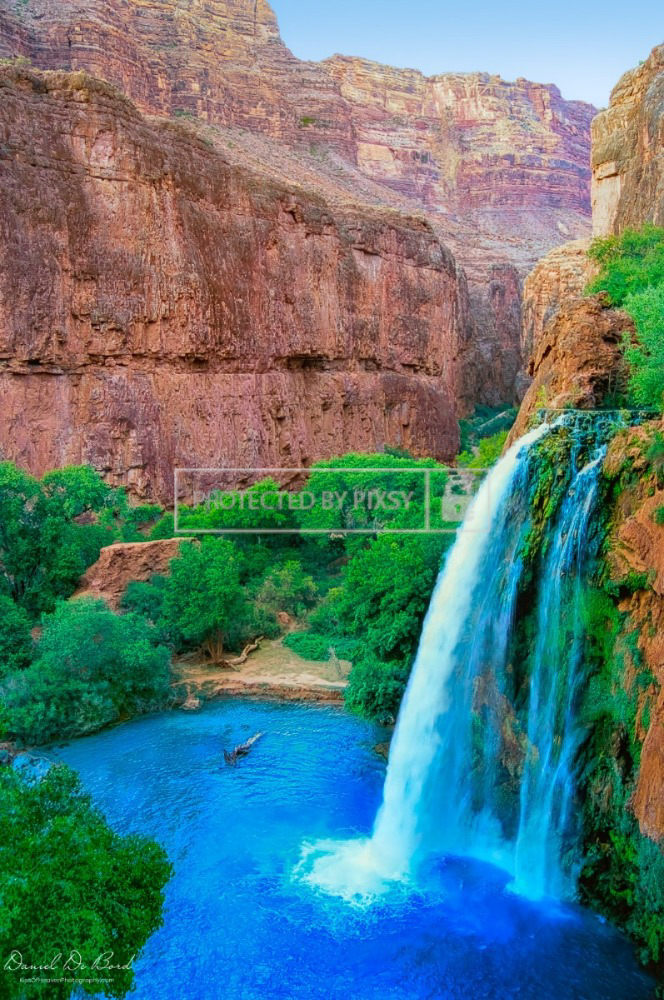 Fine art photo of a Grand Canyon waterfall flowing into a vivid blue pool, framed by red rocks under bright midday light.