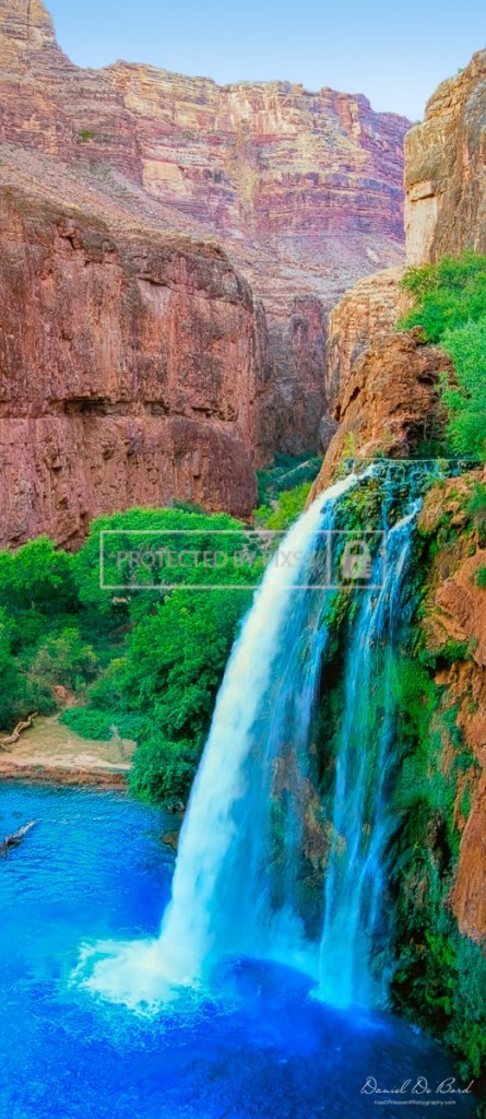 Panoramic fine art photograph of a Grand Canyon waterfall with water flowing over red rocks, surrounded by green bushes and vibrant desert scenery.