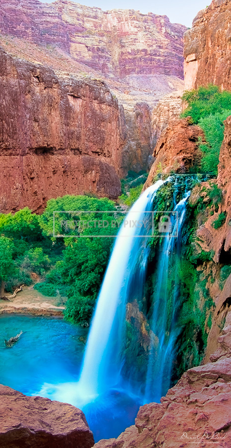 Panoramic fine art photograph of a Grand Canyon waterfall with silky water flowing over red rocks, surrounded by green bushes and vibrant desert scenery.