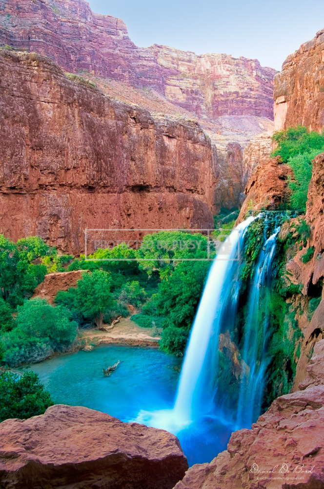 Fine art photograph of a Grand Canyon waterfall with silky water flowing over red rocks, surrounded by green bushes and vibrant desert scenery.